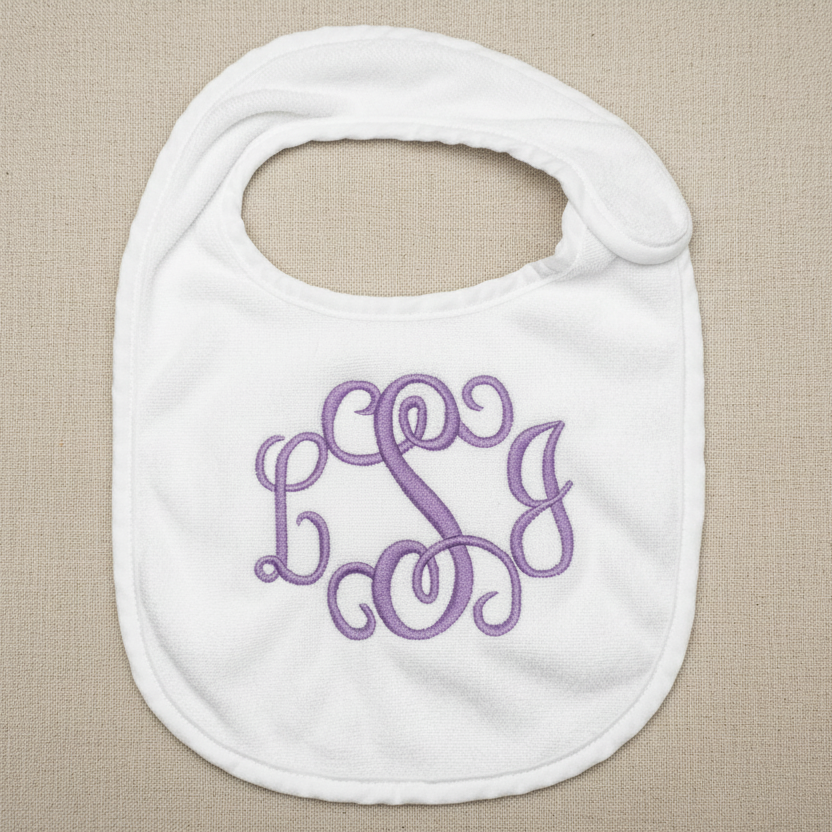 A white baby bib with a purple monogram embroidery featuring the letters 'B', 'S', and 'G' in a cursive script, displayed on a white fabric background.
