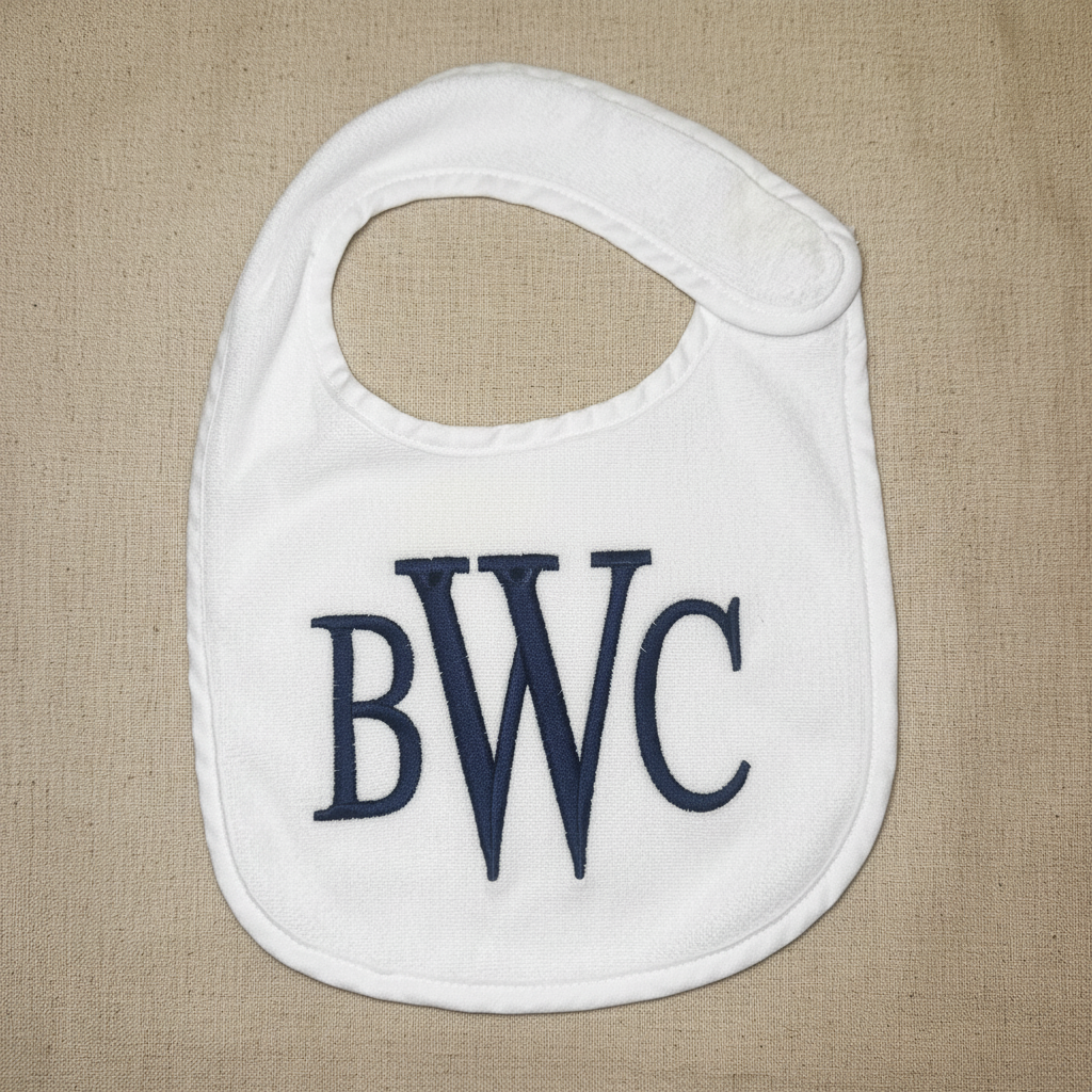 A white baby bib with navy blue embroidered letters 'BWC' on the front, laid flat on a white textured background.