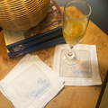 Personalized white napkins with blue embroidered text reading 'The Seranni' arranged on a wooden table, with a glass of bubbly champagne resting on one napkin and books in the background.