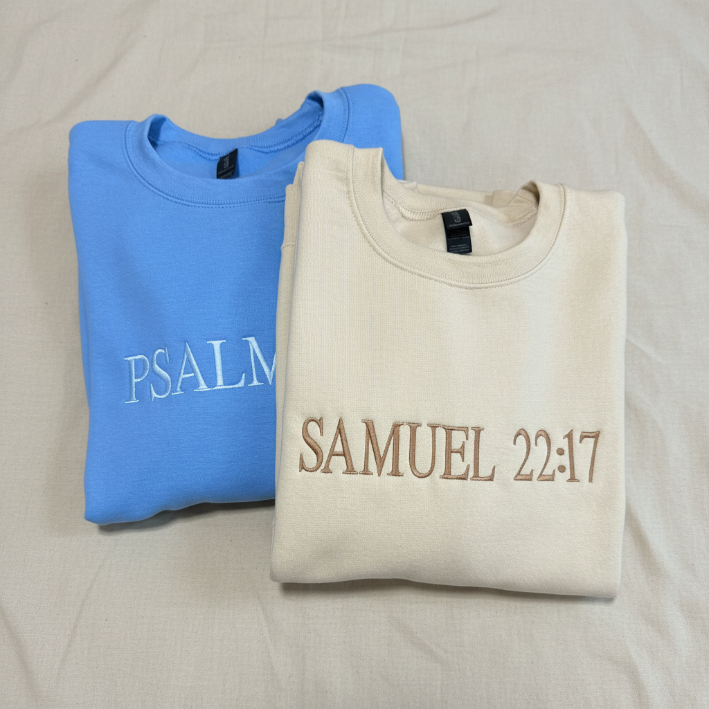 Two folded crewneck sweatshirts on a white background: one light blue with 'PSALM' embroidered in light blue, and one cream with 'SAMUEL 22:17' embroidered in brown.
