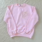 A light pink crewneck sweatshirt featuring a gold line-art graphic of three hanging bells on the left chest, laid flat against a white background.