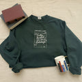 A dark forest green crewneck sweatshirt with a white graphic print of a library card that reads 'Fully Booked for the Holidays'. The sweatshirt is styled in a flat-lay arrangement on a white fabric background, accompanied by books, a mug, and a notebook.