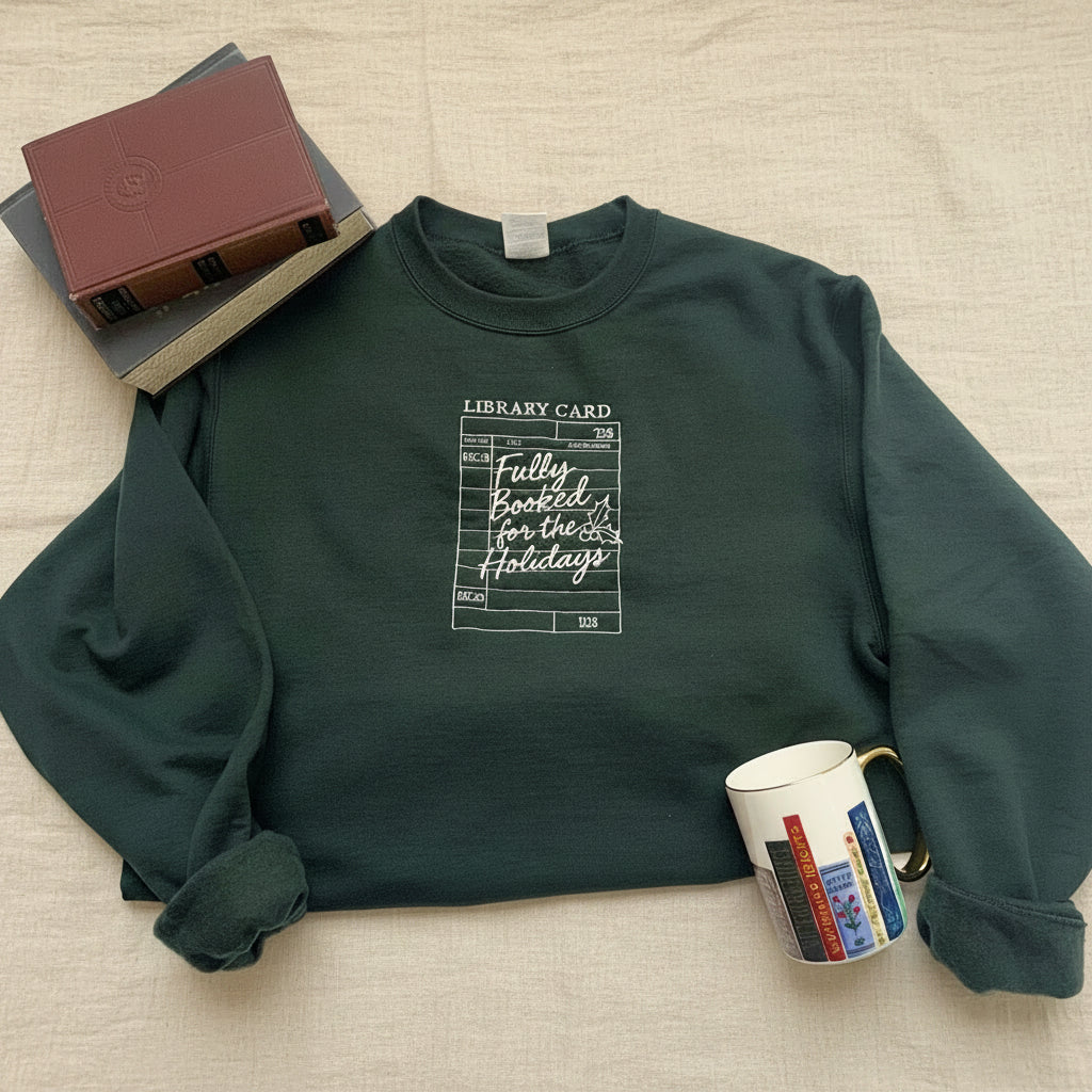 A dark forest green crewneck sweatshirt with a white graphic print of a library card that reads 'Fully Booked for the Holidays'. The sweatshirt is styled in a flat-lay arrangement on a white fabric background, accompanied by books, a mug, and a notebook.