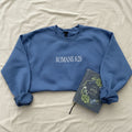 A slate blue crewneck sweatshirt with 'ROMANS 8:28' printed in white across the chest, styled in a flat lay on white fabric alongside a decorative Bible.