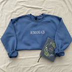 A slate blue crewneck sweatshirt with 'ROMANS 8:28' printed in white across the chest, styled in a flat lay on white fabric alongside a decorative Bible.