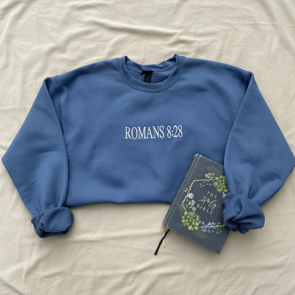 A slate blue crewneck sweatshirt with 'ROMANS 8:28' printed in white across the chest, styled in a flat lay on white fabric alongside a decorative Bible.
