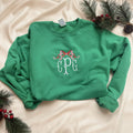 A bright green crewneck sweatshirt laid flat on white fabric, decorated with holiday pine branches, pinecones, and red berries. The front features a white and red embroidered design of three monogram letters 'C P G' under a red bow.