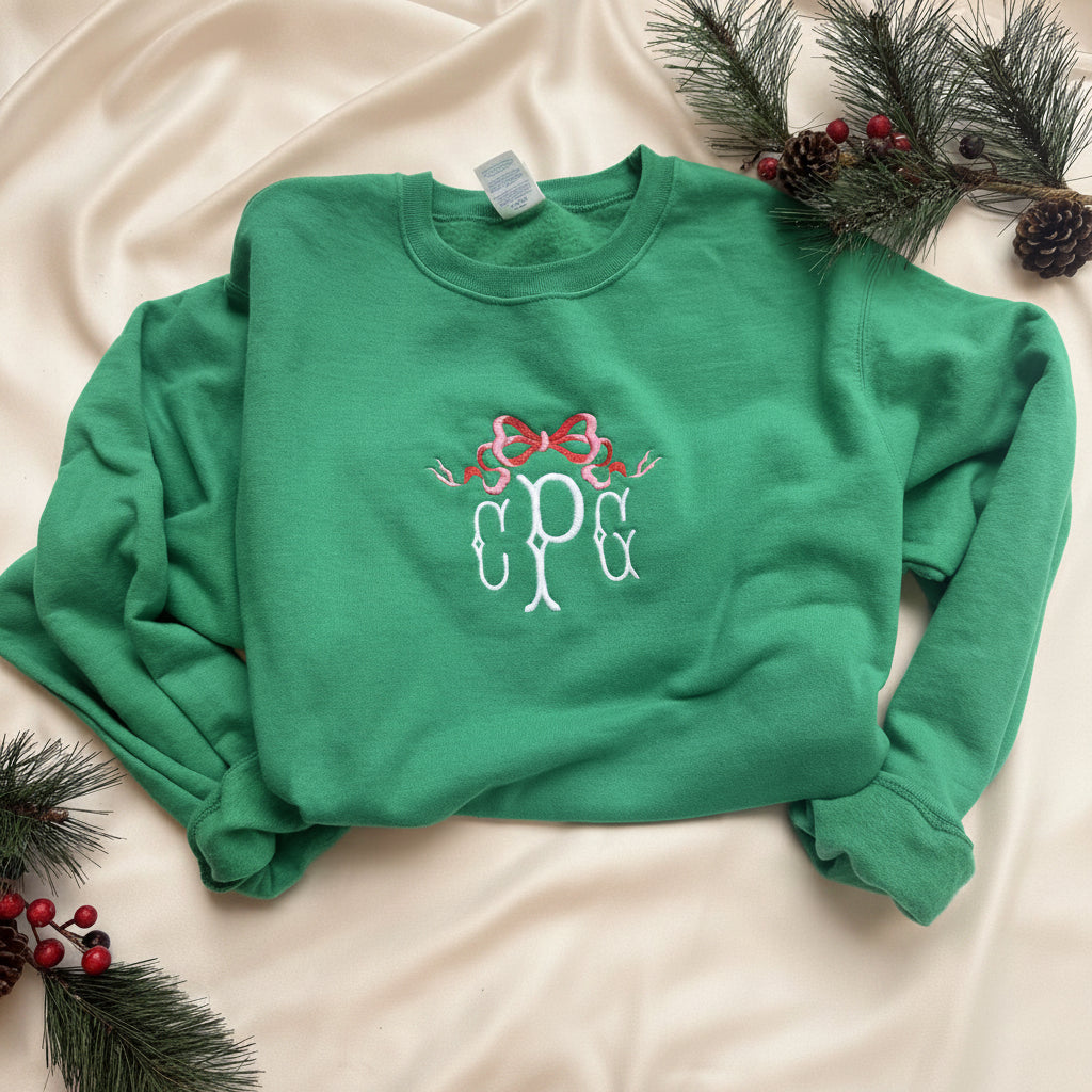 A bright green crewneck sweatshirt laid flat on white fabric, decorated with holiday pine branches, pinecones, and red berries. The front features a white and red embroidered design of three monogram letters 'C P G' under a red bow.