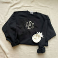 A black long-sleeve crewneck sweatshirt with a white monogram graphic on the chest, styled on a white fabric background with a decorative white flower accessory.