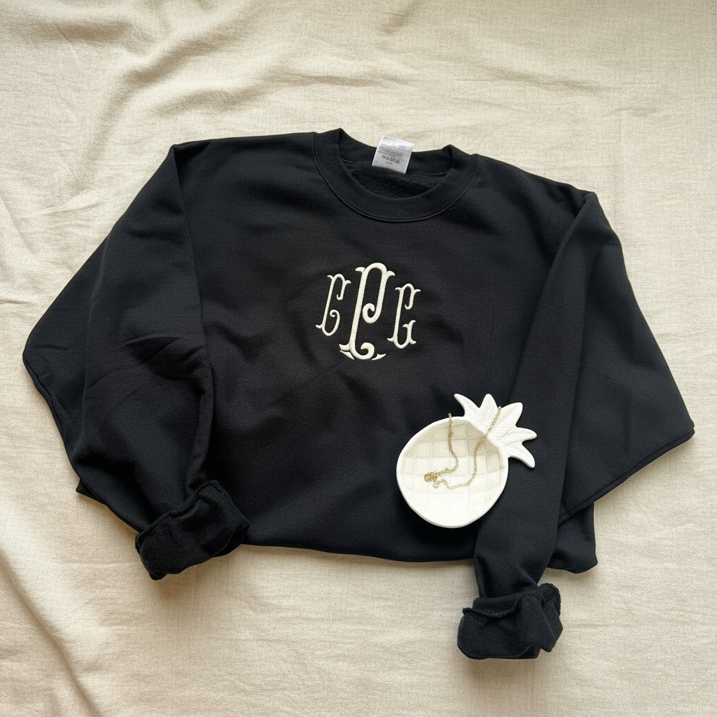 A black long-sleeve crewneck sweatshirt with a white monogram graphic on the chest, styled on a white fabric background with a decorative white flower accessory.