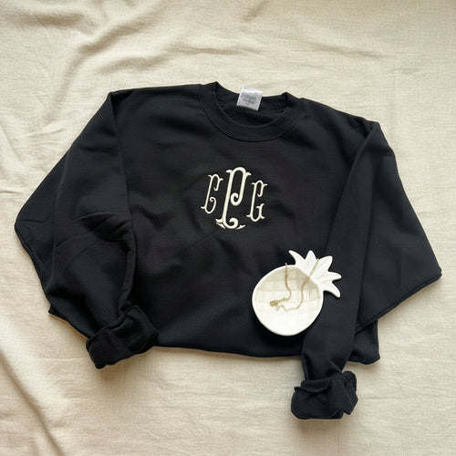 A black long-sleeve crewneck sweatshirt with a white monogram graphic on the chest, styled on a white fabric background with a decorative white flower accessory.