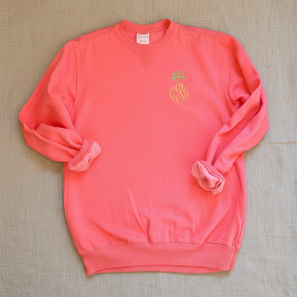 A coral pink crewneck sweatshirt with rolled-up sleeves, featuring a small gold pineapple monogram logo on the left chest. The sweatshirt is laid flat against a white background.