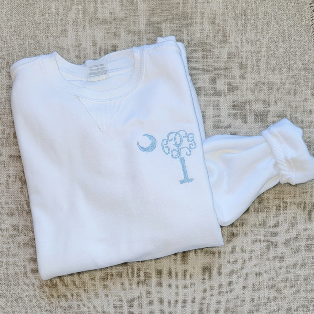 A folded white baby long-sleeve shirt with a matching footed sleeper, featuring a light blue embroidered monogram design on the chest.