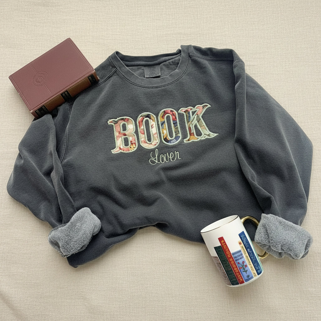 A dark gray crewneck sweatshirt with a fleece-lined collar, featuring 'BOOK Lover' printed on the front. The word 'BOOK' is styled with a colorful book-cover pattern, and 'Lover' is written in a cursive font.