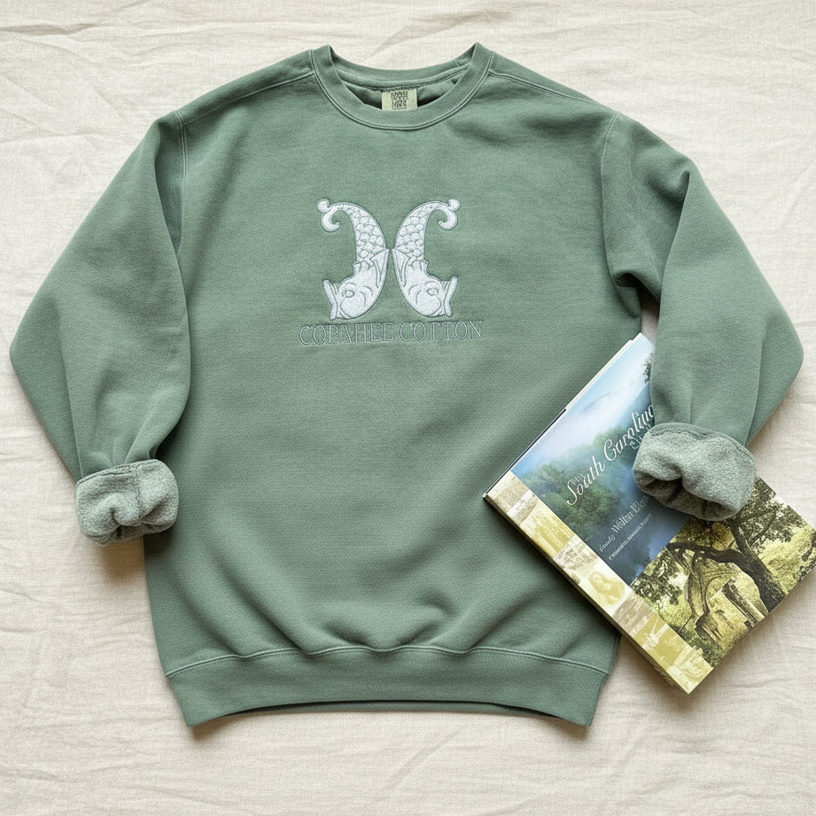 A sage green crewneck sweatshirt with a white embroidered design of two stylized pears and the text 'COPAHEE COTTON' on the chest, styled on a white fabric background next to a book.