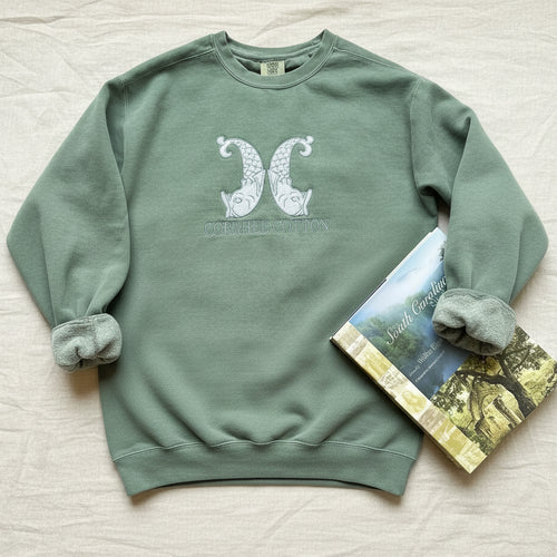 A sage green crewneck sweatshirt with a white embroidered design of two stylized pears and the text 'COPAHEE COTTON' on the chest, styled on a white fabric background next to a book.