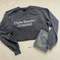 A dark gray washed crewneck sweatshirt with the phrase 'Make Heaven Crowded' in white pixel-style lettering, styled flat on a white fabric background alongside a notebook.