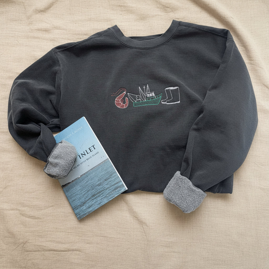 A dark charcoal gray crewneck sweatshirt with a graphic on the chest showing a shrimp, a boat, and a fish, styled on white fabric with a book and gray knit cuffs.
