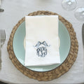 A folded white napkin with a blue embroidered monogram and bow design, placed on a light blue plate over a woven brown placemat on a beige tablecloth.