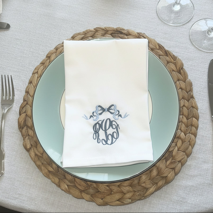 A folded white napkin with a blue embroidered monogram and bow design, placed on a light blue plate over a woven brown placemat on a beige tablecloth.