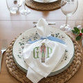 A white fabric napkin with a light blue ribbon bow and personalized embroidery of a nutcracker and the letters 'GIP', displayed on a decorative plate over a woven placemat.