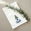 A white personalized cloth napkin with an embroidered green Christmas tree, blue pot, and the name 'Smith' in blue script, styled on a gray scalloped plate with a rosemary sprig.