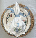 A white embroidered napkin with a blue monogram letter 'P' and floral detailing, tied with a light blue ribbon, displayed on a decorative ceramic plate over a woven placemat.