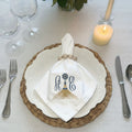 A white fabric napkin with an embroidered monogram and decorative tassel motif, tied around a white embossed plate with a braided beige charger and wooden napkin ring.