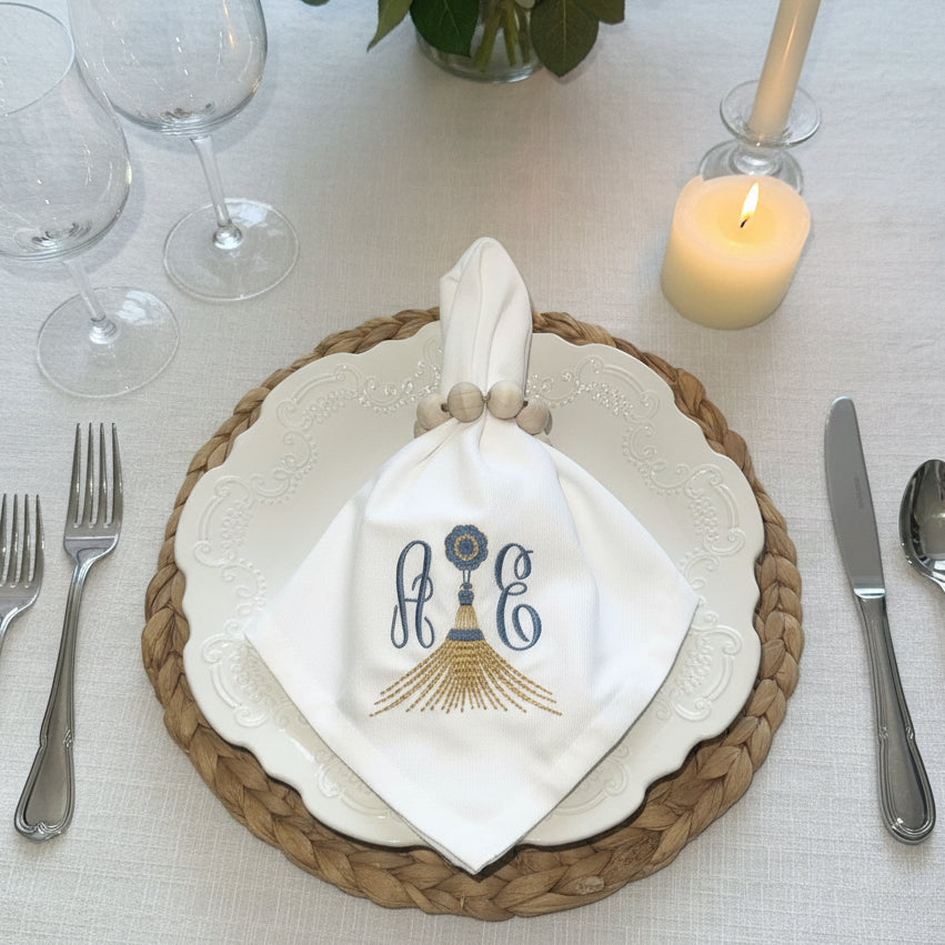 A white fabric napkin with an embroidered monogram and decorative tassel motif, tied around a white embossed plate with a braided beige charger and wooden napkin ring.