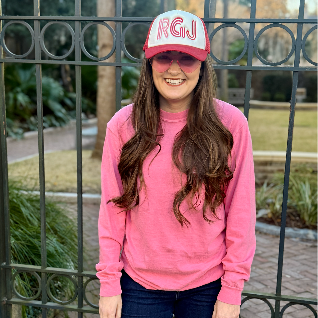 A woman standing in front of a black metal fence, wearing a bright pink crewneck sweatshirt, a pink and white trucker hat with 'RGJ' embroidered on the front, and pink sunglasses.