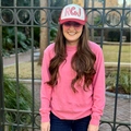A woman standing in front of a black metal fence, wearing a bright pink crewneck sweatshirt, a pink and white trucker hat with 'RGJ' embroidered on the front, and pink sunglasses.