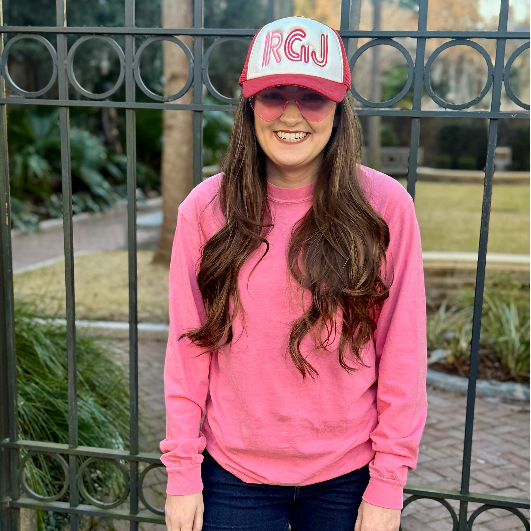 A woman standing in front of a black metal fence, wearing a bright pink crewneck sweatshirt, a pink and white trucker hat with 'RGJ' embroidered on the front, and pink sunglasses.