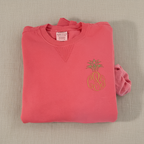 A folded coral pink crewneck sweatshirt featuring an embroidered pineapple design with a gold monogram 'CPG' on the chest, displayed on a clean white background.